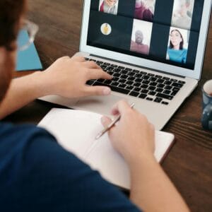 Person taking notes during a virtual meeting on a laptop screen with a diverse team.